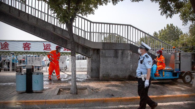 A  police officer watches as workers spray a sidewalk near the Xinfadi wholesale food market district in Beijing, Saturday, June 13, 2020. Beijing closed the city's largest wholesale food market Saturday after the discovery of seven cases of the new coronavirus in the previous two days. (AP Photo/Mark Schiefelbein)