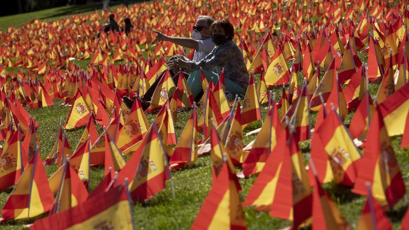 A woman sits among the Spanish flags placed in memory of coronavirus (COVID-19) victims in Madrid, Spain, Sunday, Sept. 27, 2020. An association of families of coronavirus victims has planted what it says are 53,000 small Spanish flags in a Madrid park to honor the dead of the pandemic. (AP Photo/Manu Fernandez)