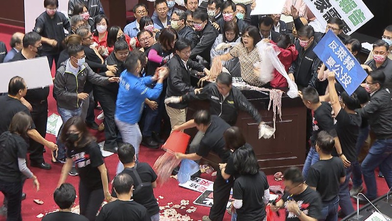 In this image made from video, Taiwanese Premier Su Tseng-chang holds a microphone as opposition party lawmakers from the Nationalist party (KMT) block his attempt to speak during a parliament session in Taipei, Taiwan, Friday, Nov. 27, 2020. Taiwan's lawmakers got into a fist fight and threw pig guts at each other Friday over a soon-to-be enacted policy that would allow imports of U.S. pork and beef. The banner at right reads: “Democratic Progressive Party feeds people with ractopamine pork.” (FTV via AP)