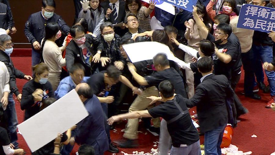 In this image made from video, Taiwanese Premier Su Tseng-chang holds a microphone as opposition party lawmakers from the Nationalist party (KMT) block his attempt to speak during a parliament session in Taipei, Taiwan, Friday, Nov. 27, 2020. Taiwan's lawmakers got into a fist fight and threw pig guts at each other Friday over a soon-to-be enacted policy that would allow imports of U.S. pork and beef. The banner at right reads: “Democratic Progressive Party feeds people with ractopamine pork.” (FTV via AP)