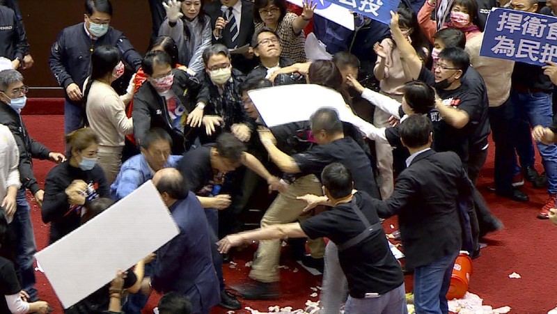 In this image made from video, Taiwanese Premier Su Tseng-chang holds a microphone as opposition party lawmakers from the Nationalist party (KMT) block his attempt to speak during a parliament session in Taipei, Taiwan, Friday, Nov. 27, 2020. Taiwan's lawmakers got into a fist fight and threw pig guts at each other Friday over a soon-to-be enacted policy that would allow imports of U.S. pork and beef. The banner at right reads: “Democratic Progressive Party feeds people with ractopamine pork.” (FTV via AP)