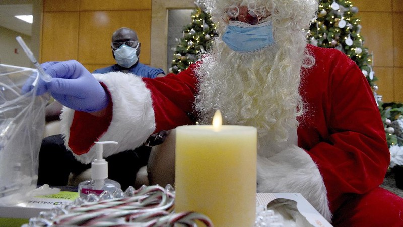 Santa, Robert Carroll, M.D., an Emergency Medicine Specialist with Manchester Memorial Hospital, takes a syringe from the plastic bag with already prepared dosages of the Pfizer-BioNTech vaccine, to be administered to Marvin Bristol, rear, a respiratory therapist and a front-line healthcare worker, Wednesday, Dec. 16, 2020, in Manchester, Conn. (Jim Michaud/Journal Inquirer via AP)