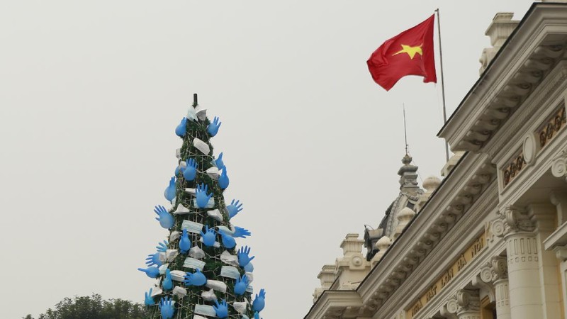 A masked security guard stands watch in front of a coronavirus-themed Christmas tree decorated with masks and rubber gloves in Hanoi, Vietnam, Thursday, Dec. 24, 2020. (AP Photo/Hau Dinh)