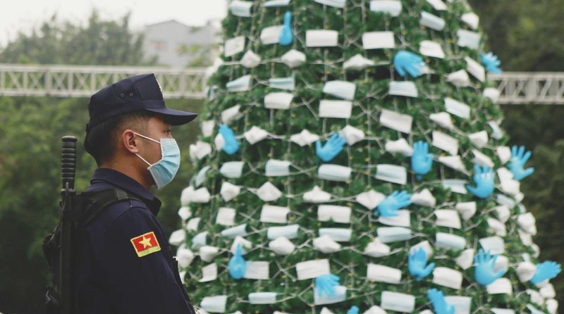 A masked security guard stands watch in front of a coronavirus-themed Christmas tree decorated with masks and rubber gloves in Hanoi, Vietnam, Thursday, Dec. 24, 2020. (AP Photo/Hau Dinh)