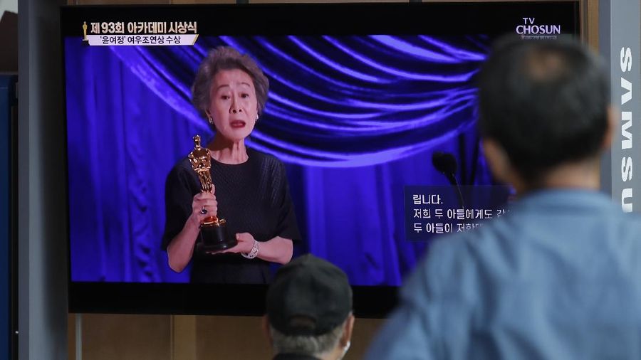 People watch live broadcast of TV showing South Korean actress Yuh-Jung Youn receiving the Oscar for best supporting actress for her performance in “Minari,” at Seoul Railway Station in Seoul, South Korea, Monday, April 26, 2021. The letters read