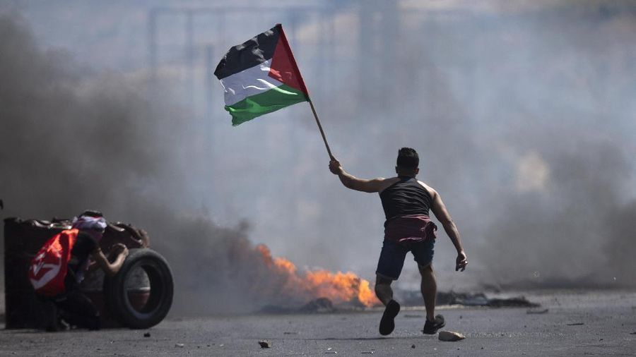 A protester waves the Palestinian flag during clashes with the Israeli forces at the Hawara checkpoint, south of the West Bank city of Nablus, Friday, May 14, 2021. Health officials say several Palestinians were killed by Israeli army fire, at protests that took place in several locations across the West Bank of Friday. (AP Photo/Majdi Mohammed)