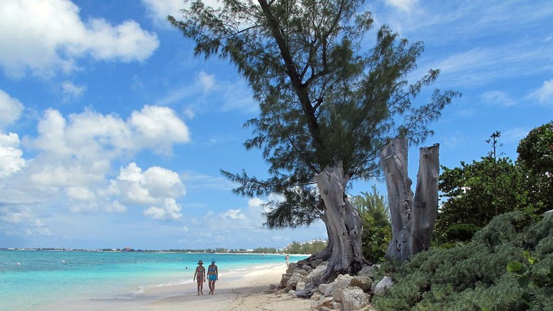 In this Aug. 3, 2012 photo, tourists walk along the beach of Seven Mile Beach in Grand Cayman Island. The Cayman Islands have lost some of their allure by abruptly proposing what amounts to an income tax on expatriate workers who have helped build the territory into one of the most famous or, for some people, notorious offshore banking centers that have tax advantages for foreign investment operations. (AP Photo/David McFadden)