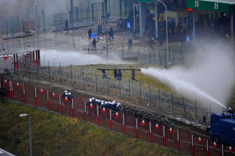 Polish law enforcement officers use water cannons, as migrants carry a log towards a fence at Kuznica - Bruzgi checkpoint, Poland, November 16, 2021. Policja Podlaska/Handout via REUTERS NO RESALES. NO ARCHIVES. MANDATORY CREDIT. THIS IMAGE HAS BEEN SUPPLIED BY A THIRD PARTY.