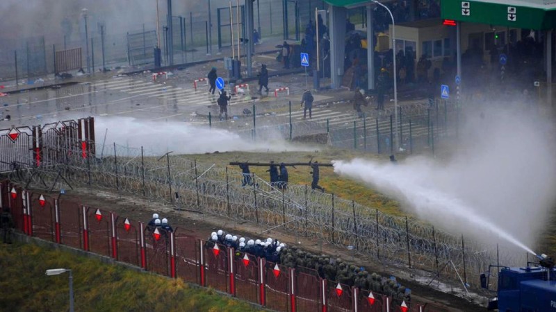 Polish law enforcement officers use water cannons, as migrants carry a log towards a fence at Kuznica - Bruzgi checkpoint, Poland, November 16, 2021. Policja Podlaska/Handout via REUTERS NO RESALES. NO ARCHIVES. MANDATORY CREDIT. THIS IMAGE HAS BEEN SUPPLIED BY A THIRD PARTY.