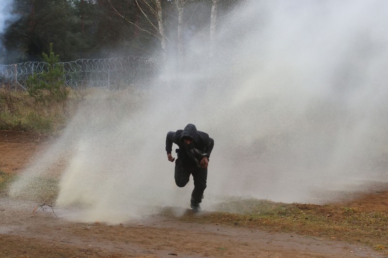 Polish law enforcement officers use water cannons, as migrants carry a log towards a fence at Kuznica - Bruzgi checkpoint, Poland, November 16, 2021. Policja Podlaska/Handout via REUTERS NO RESALES. NO ARCHIVES. MANDATORY CREDIT. THIS IMAGE HAS BEEN SUPPLIED BY A THIRD PARTY.