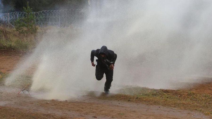 Polish law enforcement officers use water cannons, as migrants carry a log towards a fence at Kuznica - Bruzgi checkpoint, Poland, November 16, 2021. Policja Podlaska/Handout via REUTERS NO RESALES. NO ARCHIVES. MANDATORY CREDIT. THIS IMAGE HAS BEEN SUPPLIED BY A THIRD PARTY.