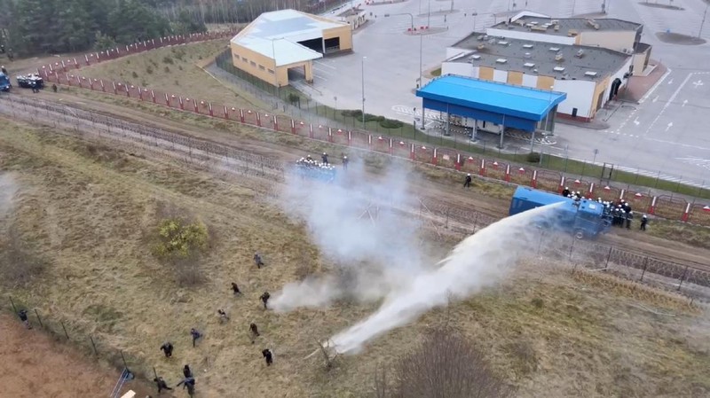 Polish law enforcement officers use water cannons, as migrants carry a log towards a fence at Kuznica - Bruzgi checkpoint, Poland, November 16, 2021. Policja Podlaska/Handout via REUTERS NO RESALES. NO ARCHIVES. MANDATORY CREDIT. THIS IMAGE HAS BEEN SUPPLIED BY A THIRD PARTY.