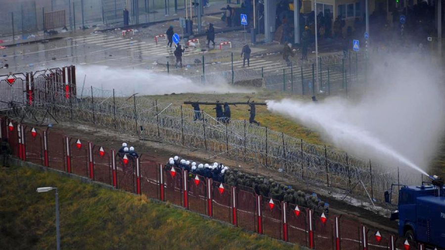 Polish law enforcement officers use water cannons, as migrants carry a log towards a fence at Kuznica - Bruzgi checkpoint, Poland, November 16, 2021. Policja Podlaska/Handout via REUTERS NO RESALES. NO ARCHIVES. MANDATORY CREDIT. THIS IMAGE HAS BEEN SUPPLIED BY A THIRD PARTY.
