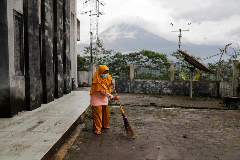Erupsi Gunung Semeru Herni, a 44-year-old local resident, sweeps ash on the ground as Mount Semeru volcano continues to spew ash and smoke in the background, at the Volcano Observatory in Candipuro district, Lumajang regency, Indonesia, December 6, 2021. REUTERS/Willy Kurniawan