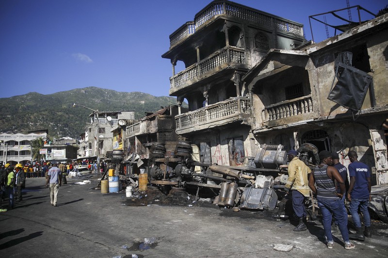 Warga berdiri di tengah rumah yang rusak akibat truk bensin terbalik dan meledak di Cap-Hatien, Haiti, Selasa (14/12/2021). Ledakan itu menghanguskan mobil dan rumah, menewaskan lebih dari 50 orang dan melukai puluhan lainnya. (AP/Joseph Odelyn)