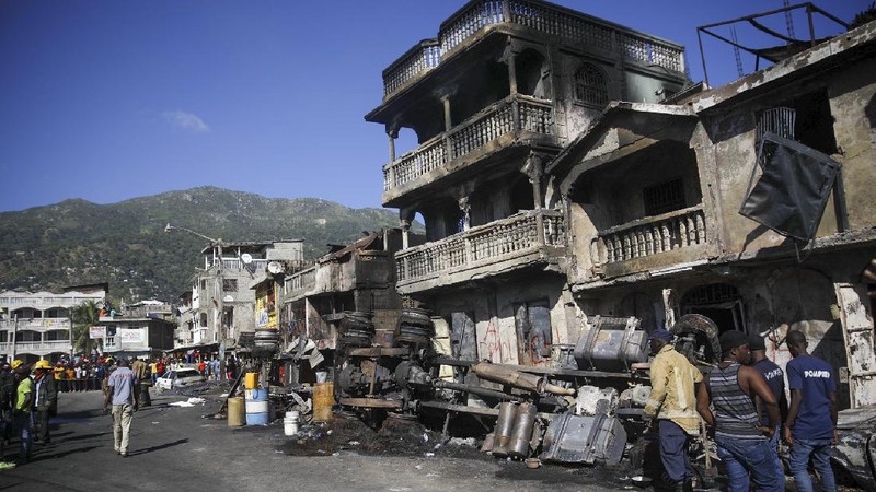 Warga berdiri di tengah rumah yang rusak akibat truk bensin terbalik dan meledak di Cap-Hatien, Haiti, Selasa (14/12/2021). Ledakan itu menghanguskan mobil dan rumah, menewaskan lebih dari 50 orang dan melukai puluhan lainnya. (AP/Joseph Odelyn)