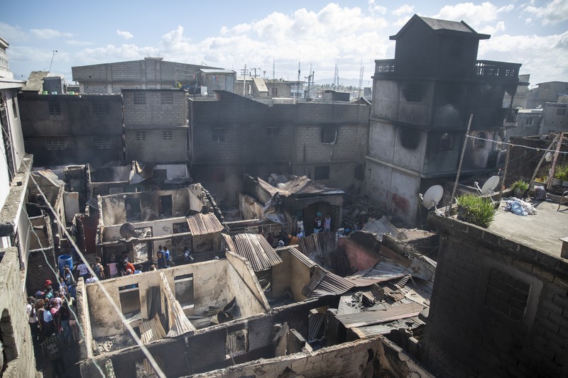 Warga berdiri di tengah rumah yang rusak akibat truk bensin terbalik dan meledak di Cap-Hatien, Haiti, Selasa (14/12/2021). Ledakan itu menghanguskan mobil dan rumah, menewaskan lebih dari 50 orang dan melukai puluhan lainnya. (AP/Joseph Odelyn)