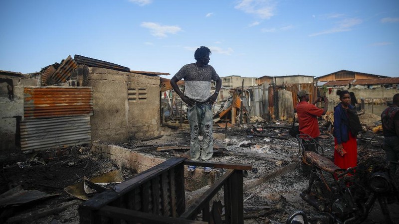 Warga berdiri di tengah rumah yang rusak akibat truk bensin terbalik dan meledak di Cap-Hatien, Haiti, Selasa (14/12/2021). Ledakan itu menghanguskan mobil dan rumah, menewaskan lebih dari 50 orang dan melukai puluhan lainnya. (AP/Joseph Odelyn)