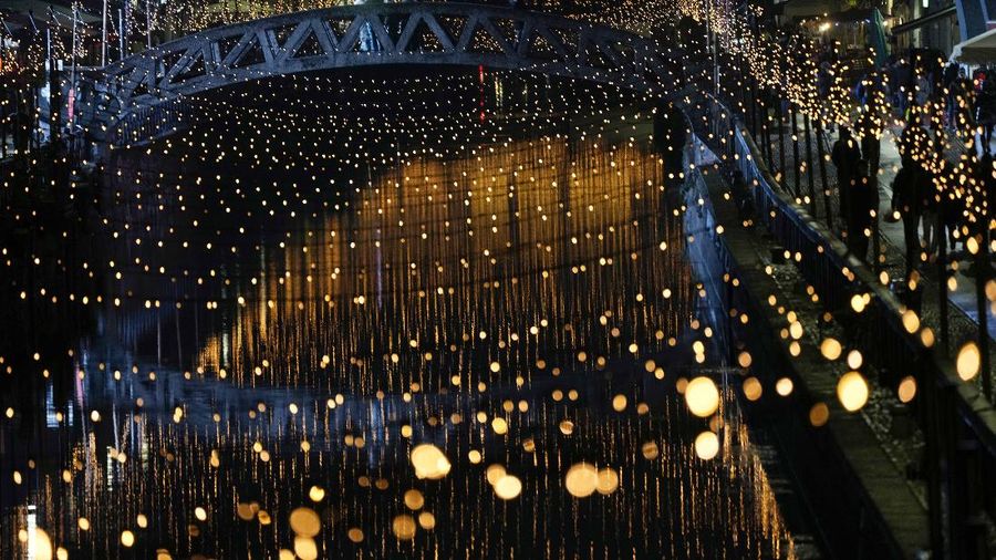 Orang-orang berjalan di jembatan saat lampu Natal menerangi Darsena dei Navigli, lingkungan yang dinamai kanal yang melintasi area Milan, Italia. (AP Photo/Luca Bruno)