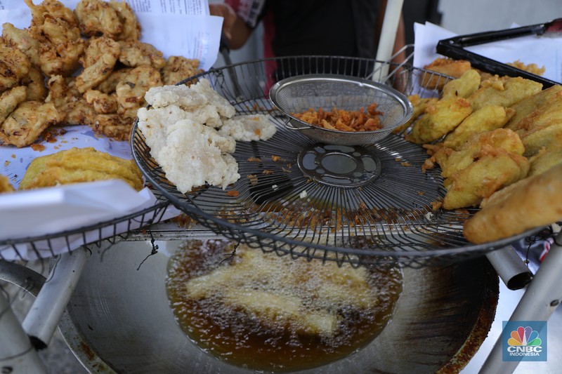 Ira Dijaya (41)melayani pembeli gorengan di kawasan Kebon Jeruk, Jakarta, Jumat (7/1/2022). Berdasarkan Pusat Informasi Harga Pangan Strategis Nasional (PIHPS), secara nasional harga minyak goreng curah pada 29 Desember lalu hanya Rp.18.400/Kg. Pada 6 Januari 2022 atau kemarin, menyentuh Rp. 18.550/Kg, atau naik 0,81%. Kemudian minyak goreng kemasan bermerk 1 pada 30 Desember 2021 harganya Rp. 20.600/Kg. Sementara kemarin menjadi Rp. 20.800/Kg, naik 0,97%. Begitu juga minyak goreng kemasan bermerk 2. Di mana pada 30 Desember masih Rp. 20.030/Kg, kemarin menjadi Rp. 20.300/Kg atau meningkat 1,34%. Harga minyak goreng di pasar tradisional masih tinggi. Pedagang menginginkan harga segera normal kembali. Menurut Ira penjual gorengan 