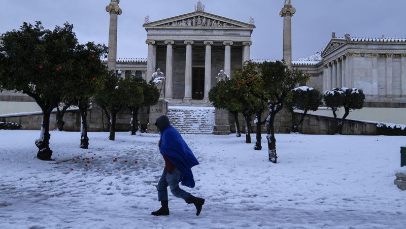 Seorang pria tunawisma dengan menggunakan seimut berjalan di atas salju di luar Akademi Athena, Selasa (25/1/2022).  Badai salju dengan tingkat keparahan yang jarang mengganggu lalu lintas jalan dan udara pada hari Senin di ibu kota Yunani Athena dan kota tetangga terbesar di Turki, Istanbul , sementara sebagian besar Yunani, termasuk — luar biasa — beberapa pulau Aegea, dan sebagian besar Turki diselimuti salju. (AP Photo/Thanassis Stavrakis)