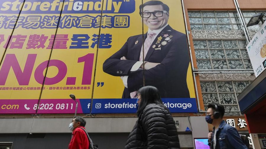 People wearing face masks walk past a huge advertisement banner in Hong Kong, Thursday, Feb. 10, 2022. Hong Kong leader Carrie Lam announced on Tuesday that places of worship and hair salons must close from Thursday until at least Feb. 24, when a “vaccine pass” will be rolled out that permits only vaccinated people to visit venues such as shopping malls and supermarkets. (AP Photo/Vincent Yu)