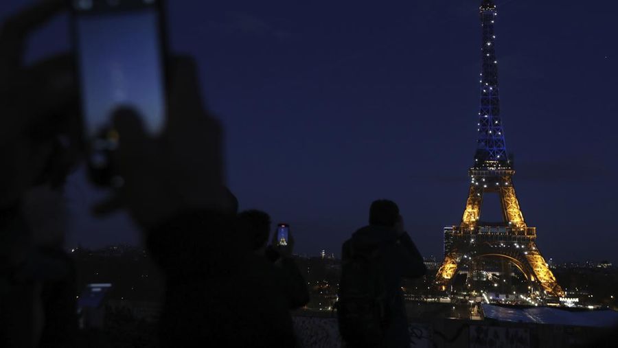 Orang-orang memotret Menara Eiffel yang diterangi dengan warna bendera Ukraina di Paris, Jumat (25/2/2022). (AP Photo/Adrienne Surprenant)