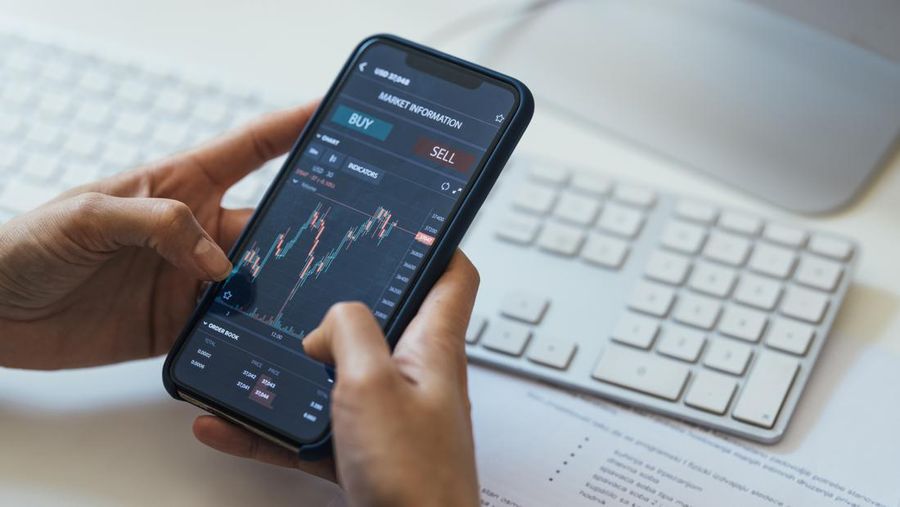 Hands of an unrecognisable woman with mobile phone on a desk, next to a keyboard, with a stock market chart on the screen