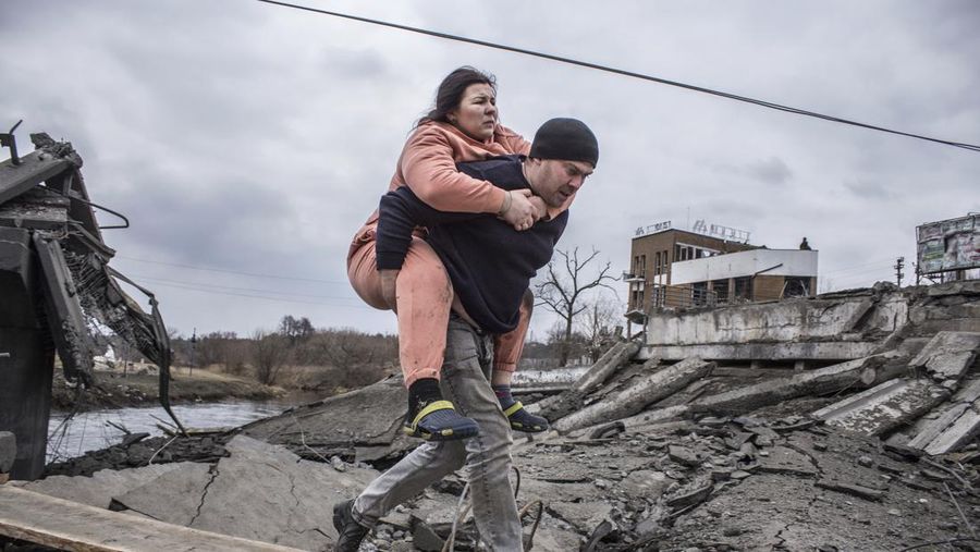 Seorang pria menggendong wanita saat mereka melintasi jalan saat melarikan diri dari kota Irpin, Ukraina, MInggu (6/3/2022). Di Irpin, dekat Kyiv, ratusan warga berjalan kaki dan bahkan dengan gerobak dorong melewati sisa-sisa jembatan yang hancur untuk menyeberangi sungai dan meninggalkan kota. (AP Photo/Oleksandr Ratushniak)