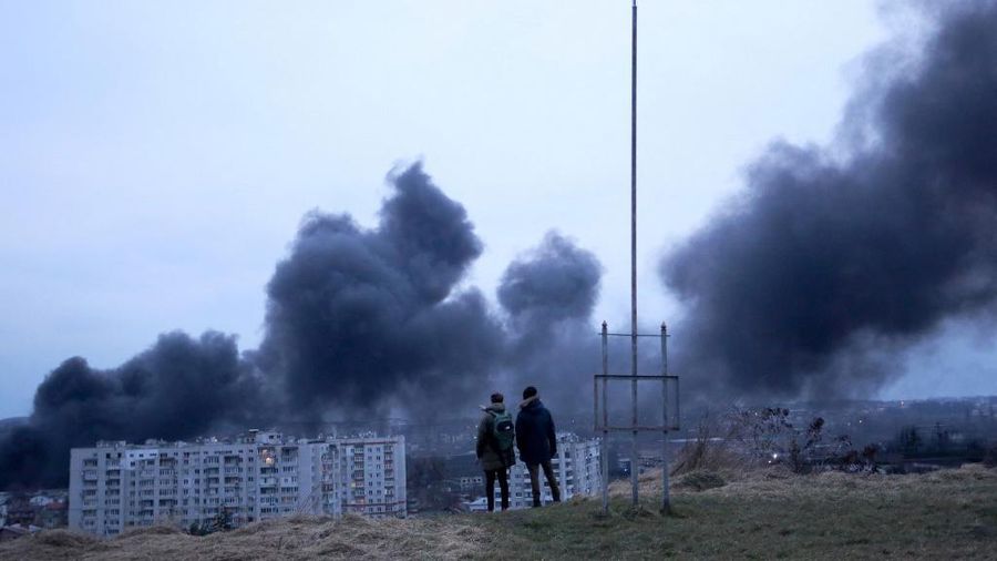 Orang-orang berdiri di depan bendera nasional Ukraina yang berkibar sambil menyaksikan asap hitam dan api membubung dari api menyusul serangan udara di kota Lviv, Ukraina barat, Sabtu (26/3/2022). Sedikitnya lima orang terluka dalam dua serangan di Lviv, kata gubernur regional, dalam serangan yang jarang terjadi di sebuah kota yang lolos dari pertempuran serius sejak pasukan Rusia menyerbu bulan lalu.  (Photo by Aleksey Filippov / AFP)