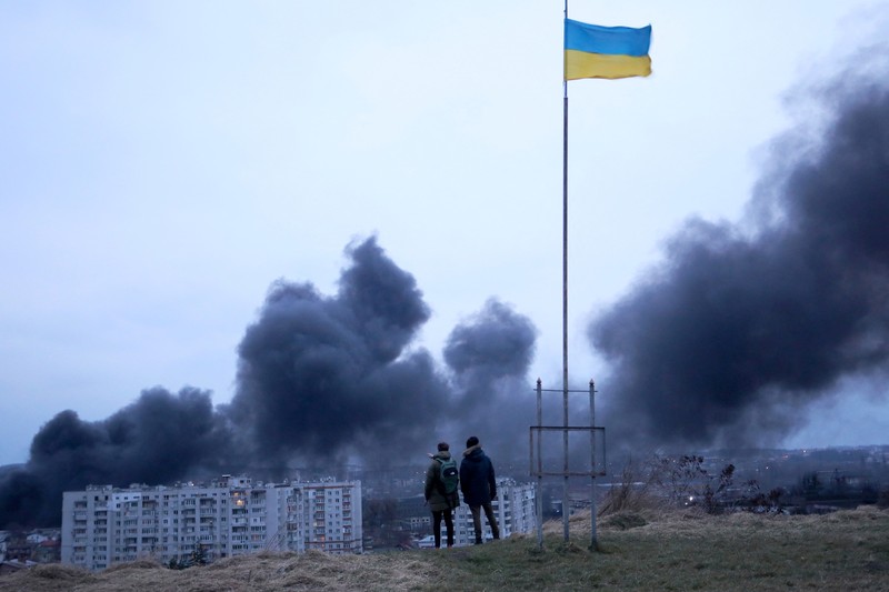 TOPSHOT - People standing near a Ukrainian national flag watch as dark smoke billows following an air strike in the western Ukrainian city of Lviv, on March 26, 2022. - At least five people wounded in two strikes on Lviv, the regional governor said, in a rare attack on a city that has escaped serious fighting since Russian troops invaded last month. (Photo by Aleksey Filippov / AFP) (Photo by ALEKSEY FILIPPOV/AFP via Getty Images)