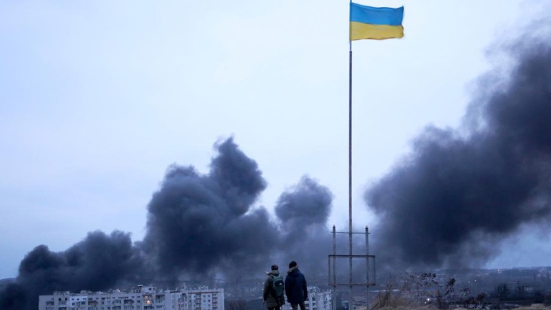 TOPSHOT - People standing near a Ukrainian national flag watch as dark smoke billows following an air strike in the western Ukrainian city of Lviv, on March 26, 2022. - At least five people wounded in two strikes on Lviv, the regional governor said, in a rare attack on a city that has escaped serious fighting since Russian troops invaded last month. (Photo by Aleksey Filippov / AFP) (Photo by ALEKSEY FILIPPOV/AFP via Getty Images)