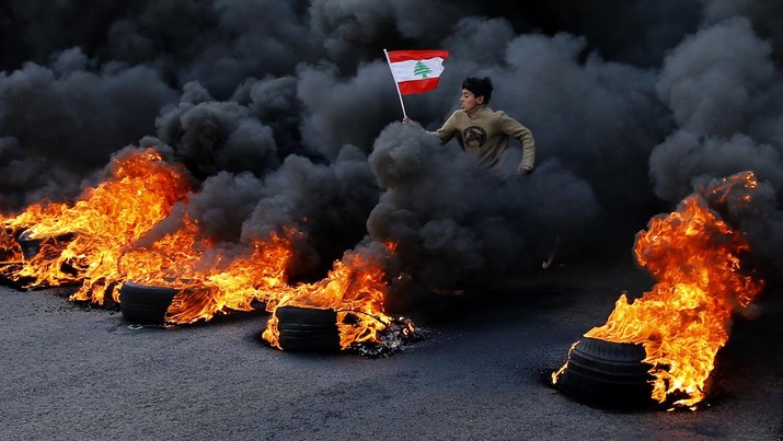 FILE- An anti-government demonstrator holds a national flag and runs across tires that were set on fire to block a main highway during a protest against a ruling elite they say has failed to address the economy's downward spiral, in the town of Jal el-Dib, north of Beirut, Lebanon, Tuesday, Jan. 14, 2020. Lebanon and Sri Lanka may be a world apart, but they share a history of political turmoil and violence that led to the collapse of once-prosperous economies bedeviled by corruption, patronage, nepotism and incompetence. (AP Photo/Bilal Hussein, File)