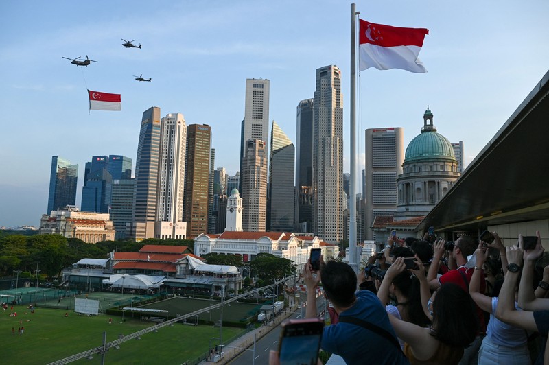 Perayaan Hari Nasional di Singapura Penonton bersorak saat helikopter Chinook Angkatan Udara Singapura dikawal oleh helikopter Apache berparade dengan bendera Singapura untuk menandai Hari Nasional ke-57 negara itu, di Singapura, Selasa (9/8/2022). (Photo by ROSLAN RAHMAN/AFP via Getty Images)