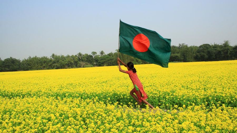 A girl runs through a mustard field hoisting the National flag. Manikganj, Bangladesh. February 5, 2008. (Photo by: Majority World/Universal Images Group via Getty Images)