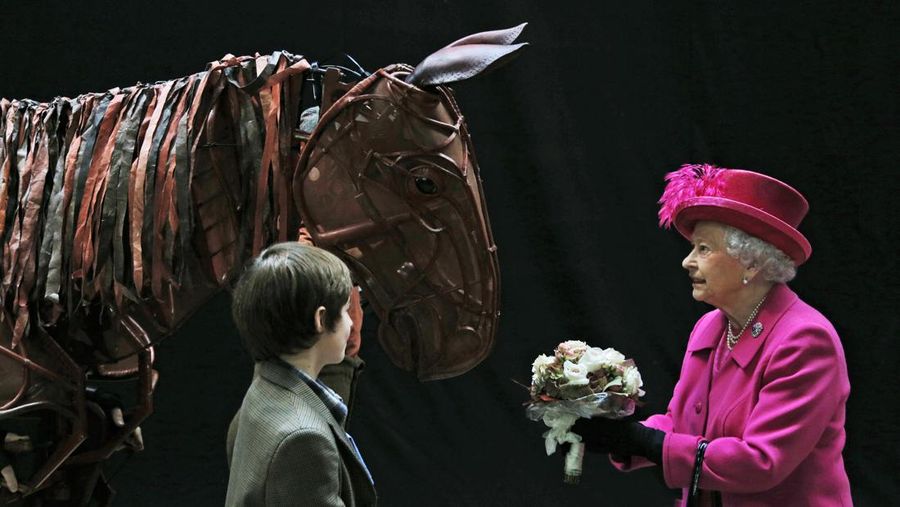 Ratu Inggris Elizabeth II menerima bunga dari seorang aktor cilik saat dia memeriksa penyangga kuda dari produksi teater 'War Horse' di National Theatre di London, 22 Oktober 2013, saat berkunjung untuk memperingati ulang tahun ke-50 institusi tersebut. (Photo by LEFTERIS PITARAKIS/POOL/AFP via Getty Images)