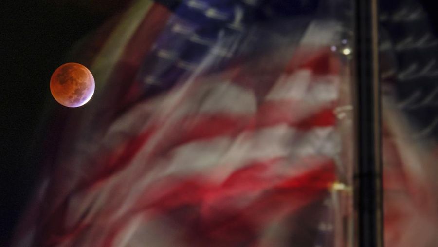 Bulan purnama akibat gerhana bulan dibingkai oleh Bendera AS yang tertiup angin di National Mall di Washington, Selasa, 8 November 2022. (AP Photo/J. David Ake)