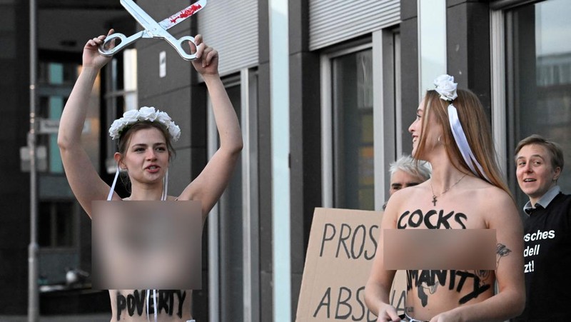 Seorang anggota kelompok aktivis feminis Femen mementaskan pertunjukan di samping seorang demonstran yang memegang poster bertuliskan 'Hapus Prostitusi' di depan gedung BPK (Bundespressekonferenz)  sebelum konferensi pers yang mempresentasikan hasil penelitian tentang prostitusi dan klien mereka di Berlin, Jerman, Selasa (8/11/2022). (Photo by TOBIAS SCHWARZ/AFP via Getty Images)