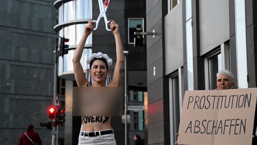 Seorang anggota kelompok aktivis feminis Femen mementaskan pertunjukan di samping seorang demonstran yang memegang poster bertuliskan 'Hapus Prostitusi' di depan gedung BPK (Bundespressekonferenz)  sebelum konferensi pers yang mempresentasikan hasil penelitian tentang prostitusi dan klien mereka di Berlin, Jerman, Selasa (8/11/2022). (Photo by TOBIAS SCHWARZ/AFP via Getty Images)