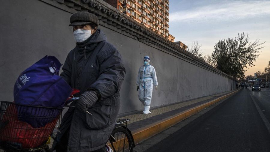 BEIJING, CHINA - DECEMBER 04: A woman wears a  mask as she rides by an epidemic control worker wearing PPE as he walks in the street near a community with residents under health monitoring for COVID-19 on December 4, 2022 in Beijing, China. In recent weeks, China has been recording some of its highest number of COVID-19 cases since the pandemic began, while some restrictions have been relaxed, authorities are sticking to their approach to containing the virus with targeted lockdowns and testing, mask mandates, and quarantines. The government has launched a vaccination drive targeting the elderly, and indicated that some positive cases may be able to quarantine at home instead of a government facility. (Photo by Kevin Frayer/Getty Images)