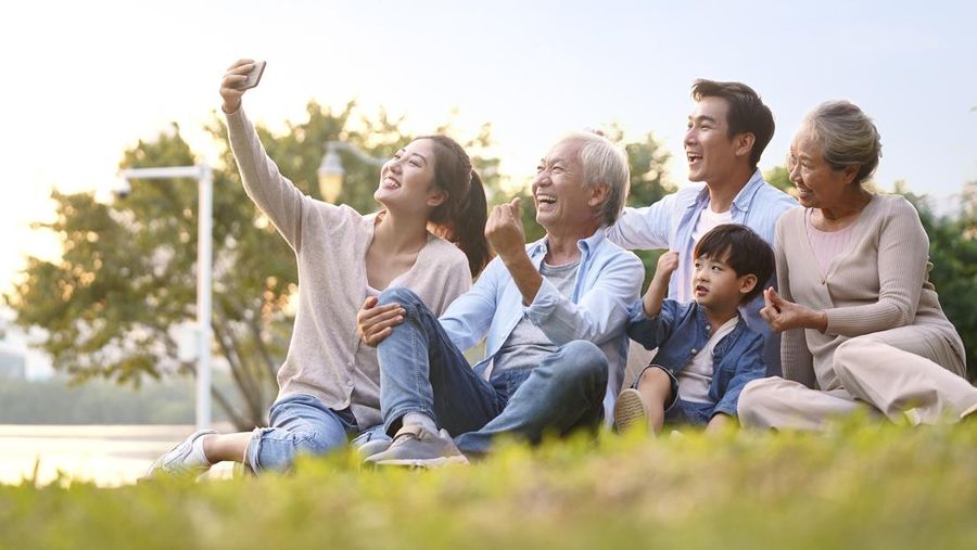 three generation happy asian family sitting on grass taking a selfie using mobile phone outdoors in park
