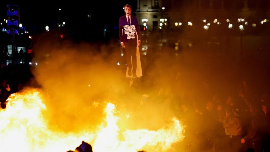 Seorang pengunjuk rasa memegang gambar Presiden Prancis Emmanuel Macron di dekat api selama demonstrasi di Place de la Concorde untuk memprotes penggunaan pasal 49.3 oleh pemerintah Prancis, klausul khusus dalam Konstitusi Prancis, untuk mendorong RUU reformasi pensiun melalui Majelis Nasional tanpa pemungutan suara oleh anggota parlemen, di Paris, Prancis, 17 Maret 2023. (REUTERS/Gonzalo Fuentes)