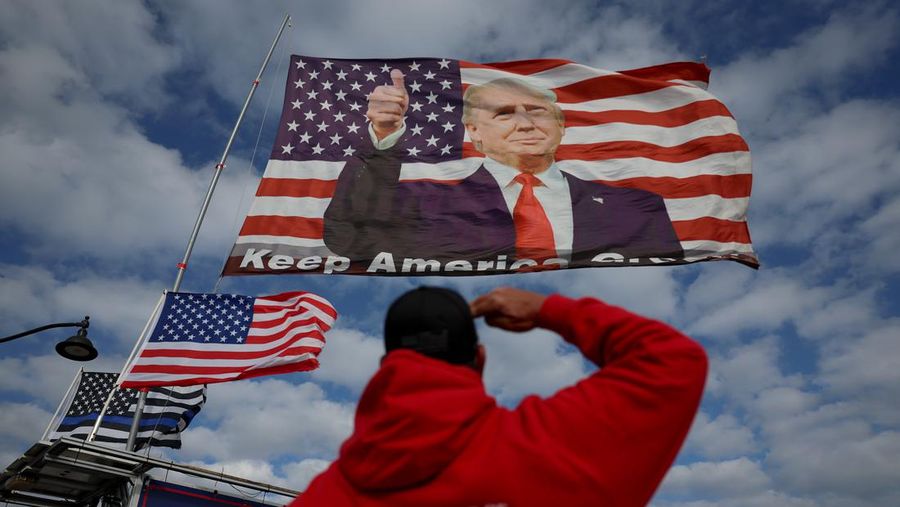 Derek Arnold memberi hormat pada bendera yang menampilkan mantan Presiden Donald Trump yang dikibarkan pendukungnya di dekat rumahnya di Mar-a-Lago pada 20 Maret 2023 di Palm Beach, Florida. Trump mengatakan di sebuah unggahan media sosial bahwa dia memperkirakan akan ditangkap sehubungan dengan penyelidikan skema uang tutup mulut yang melibatkan aktris film dewasa Stormy Daniels dan meminta para pendukungnya untuk memprotes tindakan tersebut. Namun, belum jelas apakah dia akan ditangkap atau tidak. (Joe Raedle/Getty Images)