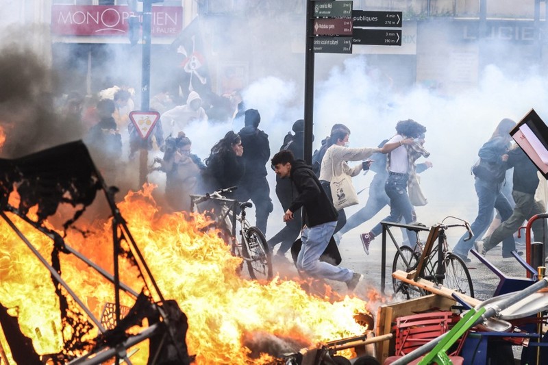 Protesters run in tear gas smoke next to   a street fire on the sidelines of a demonstration as part of a national day of strikes and protests, a week after the French government pushed a pensions reform through parliament without a vote, using the article 49.3 of the constitution, in Toulouse, southern France, on March 23, 2023. - French unions on March 23 staged a new day of disruption against the president's pension reform after he defiantly vowed to implement the change, which includes raising the age of retirement from 62 to 64, saying he was prepared to accept unpopularity in the face of sometimes violent protests. (Photo by Charly TRIBALLEAU / AFP)