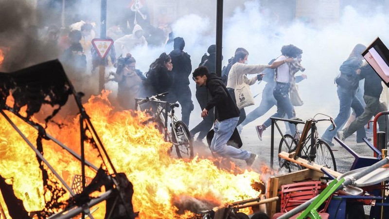 Protesters run in tear gas smoke next to   a street fire on the sidelines of a demonstration as part of a national day of strikes and protests, a week after the French government pushed a pensions reform through parliament without a vote, using the article 49.3 of the constitution, in Toulouse, southern France, on March 23, 2023. - French unions on March 23 staged a new day of disruption against the president's pension reform after he defiantly vowed to implement the change, which includes raising the age of retirement from 62 to 64, saying he was prepared to accept unpopularity in the face of sometimes violent protests. (Photo by Charly TRIBALLEAU / AFP)