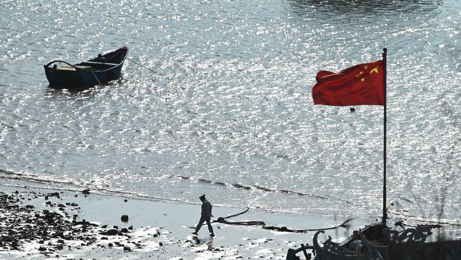 Seorang pria berjalan di pantai dekat bendera China di pulau Pingtan, di seberang Taiwan, di provinsi Fujian tenggara China pada 9 April 2023. - China sedang melakukan latihan militer hari kedua di sekitar Taiwan pada 9 April, yang disebutnya sebagai 