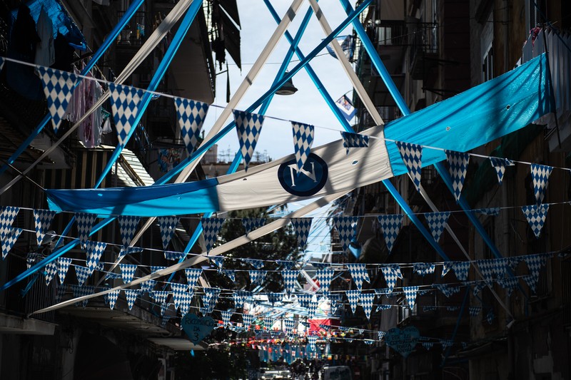 Bendera, spanduk, dan pita dipajang di jalan-jalan untuk merayakan kemenangan gelar Serie A ketiga pada 27 April 2023 di Naples, Italia. Kota Naples sedang bersiap untuk merayakan kemenangan Scudetto ketiga di kejuaraan Serie A. Di jalan-jalan pusat bersejarah dan di berbagai distrik Napoli, dekorasi, bendera, dan spanduk telah dipasang untuk merayakan kejuaraan sepak bola yang dimainkan oleh tim yang dilatih oleh Spalletti. Para pemain bisa menjadi juara Italia lebih awal, jika mereka berhasil mengalahkan Salernitana dalam derby Campania dalam pertandingan yang akan dimainkan pada hari Minggu pukul 15:00 di Stadion Diego Armando Maradona dan jika Lazio gagal menang tandang melawan Internazionale. Napoli telah memenangkan dua Scudetti dalam sejarahnya pada musim 1986-1987 dan 1989-1990, kedua kemenangan tersebut diperoleh dengan kehadiran Diego Armando Maradona, para fans Neapolitan menunggu untuk dapat merayakan 33 tahun setelah kesuksesan terakhir. (Ivan Romano/Getty Images)