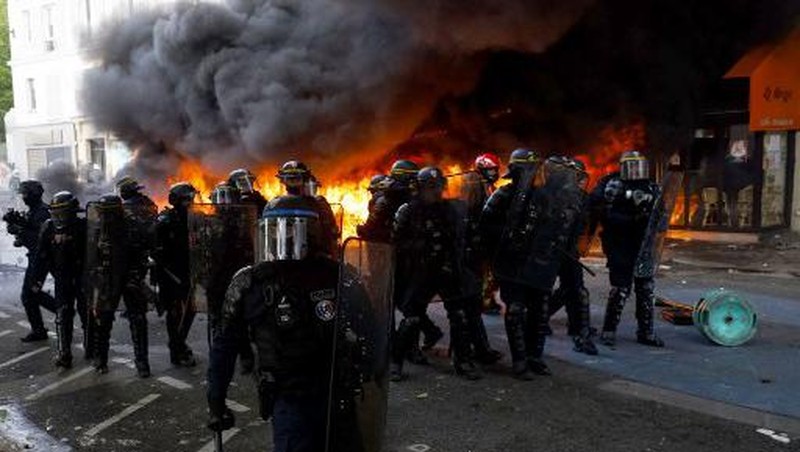 French Republican Security Corps (CRS - Compagnies Republicaines de Securite) police officers and Police officers take position next to a burning bicycle-sharing station during a demonstration on May Day (Labour Day), to mark the international day of the workers, more than a month after the government pushed an unpopular pensions reform act through parliament, in Paris, on May 1, 2023. - Opposition parties and trade unions have urged protesters to maintain their three-month campaign against the law that will hike the retirement age to 64 from 62. (Photo by Geoffroy Van der Hasselt / AFP)