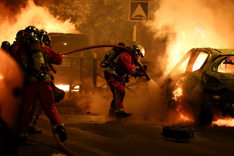 Petugas pemadam kebakaran berdiri saat mereka memadamkan kendaraan yang terbakar selama bentrokan antara pengunjuk rasa dan polisi, setelah kematian Nahel, seorang remaja berusia 17 tahun yang dibunuh oleh seorang petugas polisi Prancis saat menghentikan lalu lintas, di Nanterre, pinggiran kota Paris, Prancis, 28 Juni 2023. (REUTERS/Stephanie Lecocq)