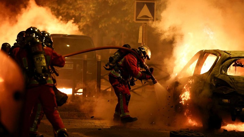 Petugas pemadam kebakaran berdiri saat mereka memadamkan kendaraan yang terbakar selama bentrokan antara pengunjuk rasa dan polisi, setelah kematian Nahel, seorang remaja berusia 17 tahun yang dibunuh oleh seorang petugas polisi Prancis saat menghentikan lalu lintas, di Nanterre, pinggiran kota Paris, Prancis, 28 Juni 2023. (REUTERS/Stephanie Lecocq)